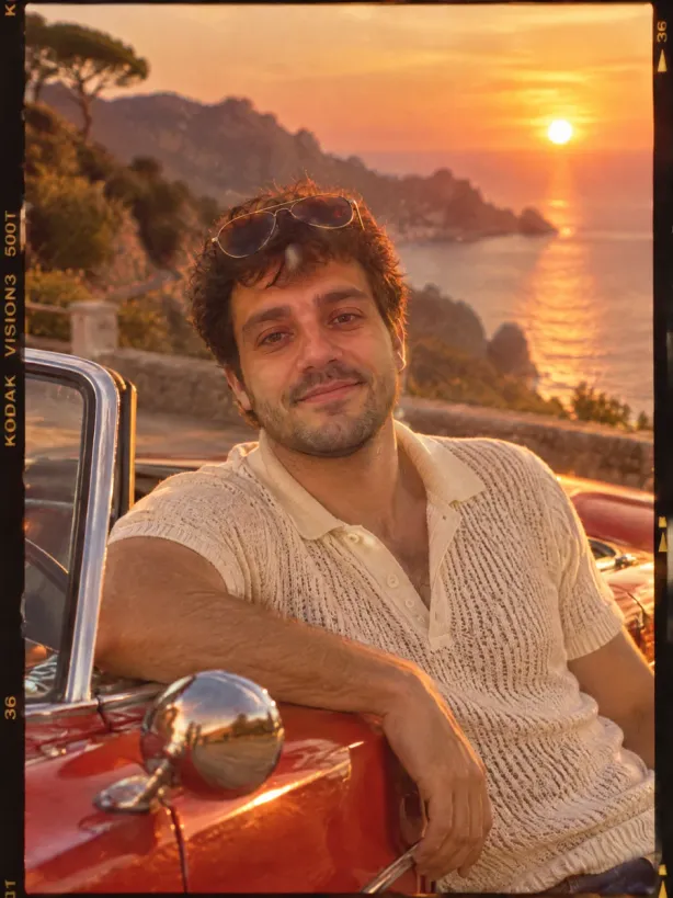 The same man in a 1970s film still next to a red convertible at sunset